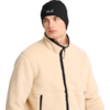 Mann, Brustbild, trägt beige Fleecejacke mit schwarzem Reißverschluss und schwarzen Taschenkanten, schwarzer Beanie mit weißem Logo, blickt nach links, weißer Hintergrund