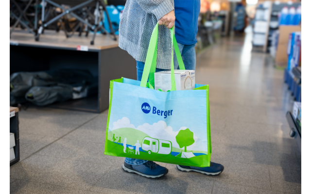 Person hält eine grün-blaue wiederverwendbare Tasche mit der Aufschrift "Berger" und einer Illustration eines Wohnwagens; sichtbare Beine und Schuhe, Ladenregale im Hintergrund