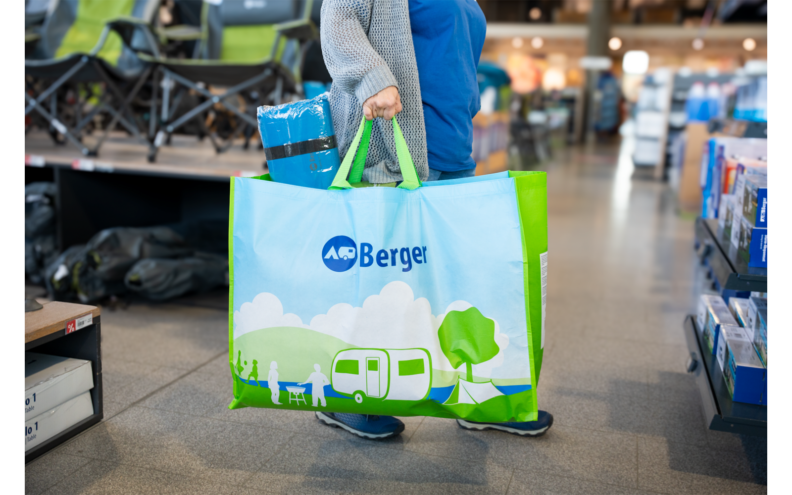 Person hält große Tasche mit grünen Henkeln und Aufdruck "Berger" (Wohnwagen, Bäume), im Hintergrund Regale und Klappstühle