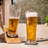 Two tall beer glasses with golden liquid and foam, one tilted in a wooden holder and one upright on a coaster on a kitchen counter by a window; basil plant to the right, hanging kitchen brush to the left