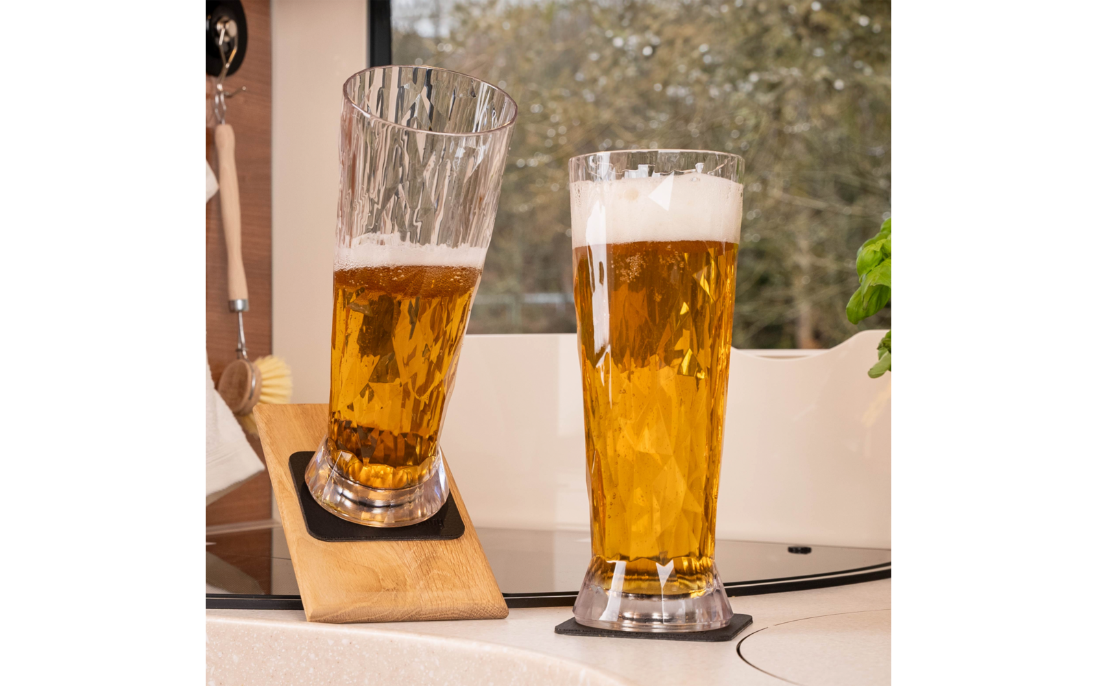 Two tall beer glasses with golden liquid and foam, one tilted in a wooden holder and one upright on a coaster on a kitchen counter by a window; basil plant to the right, hanging kitchen brush to the left