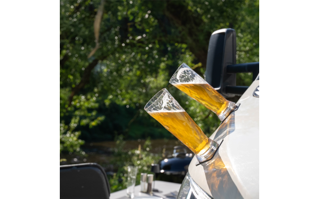 Two tilted beer glasses in holders attached to a white surface, blurred background with trees and water