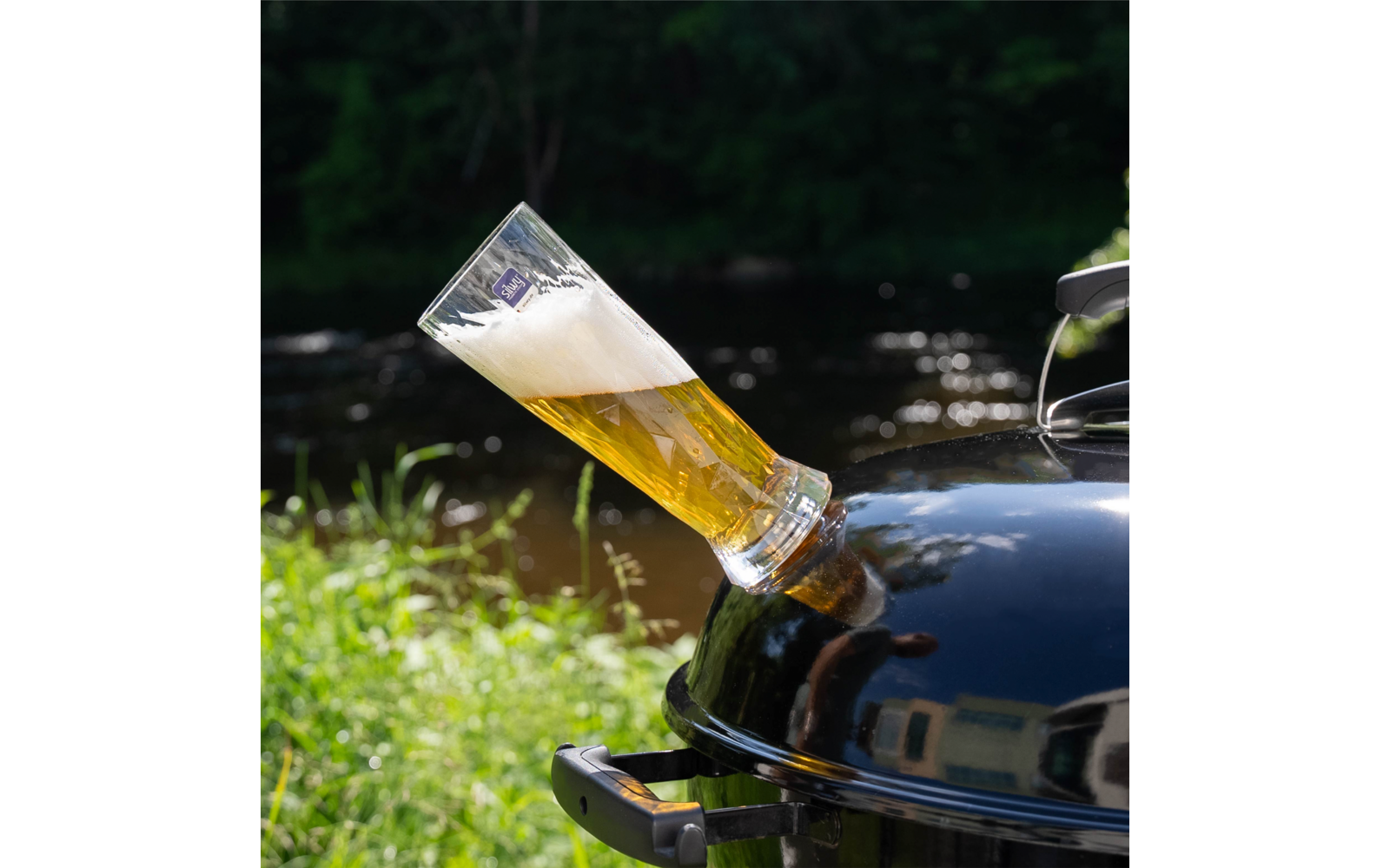 Beer glass with foam inserted into an opening of a black kettle grill; water, grass and trees in the background