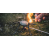 Hand die met een lucifer een kleine gasbrander op een driepoot op gras aansteekt; gasslang zichtbaar