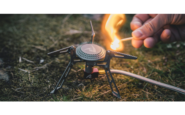 Hand die met een lucifer een kleine gasbrander op een driepoot op gras aansteekt; gasslang zichtbaar
