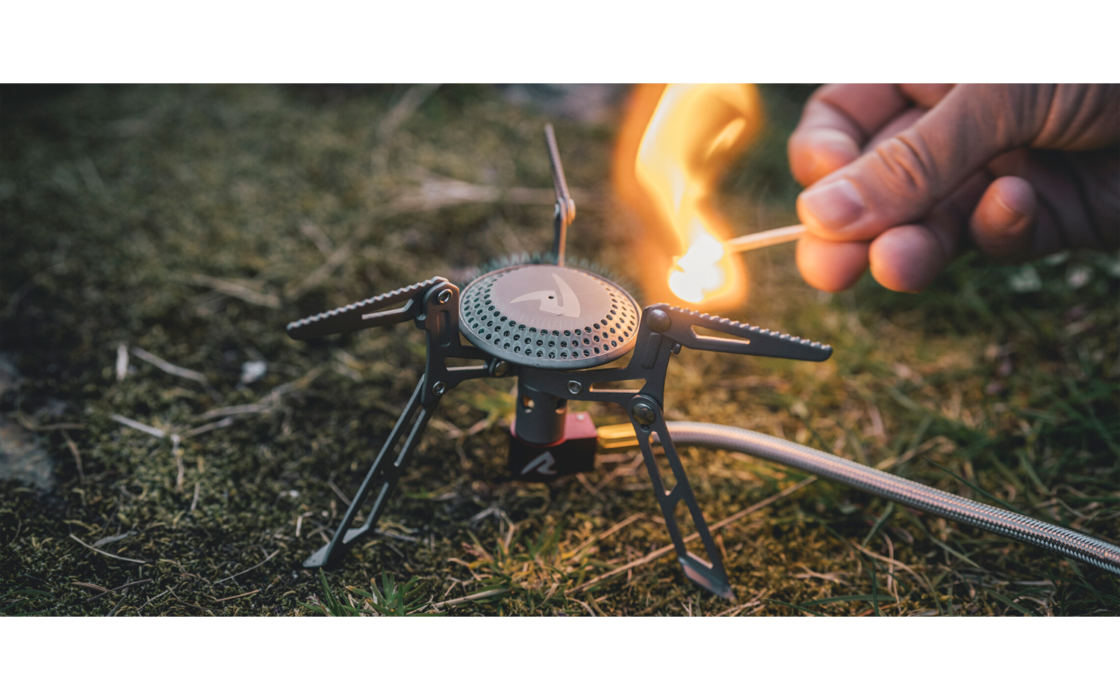 Hand die met een lucifer een kleine gasbrander op een driepoot op gras aansteekt; gasslang zichtbaar