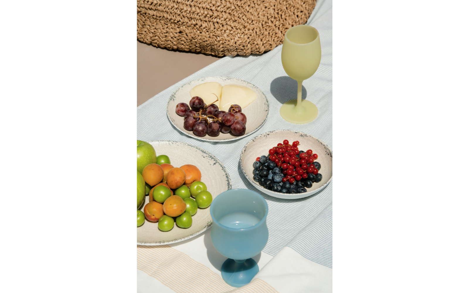 Drei Keramikschalen mit roten Trauben und hellen runden Scheiben, roten Johannisbeeren und Blaubeeren, grünen Limetten und kleinen orangen Früchten; zwei farbige Kelchgläser (gelb, blau) auf gestreiftem hellblau-beigem Tuch und geflochtenem Strohunte