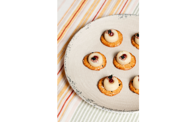 Sechs runde Cracker mit weißen Tupfen, darauf kleine rote Beeren und grüne Kräuterblättchen, auf einem beigen Teller auf einem gestreiften Tischtuch