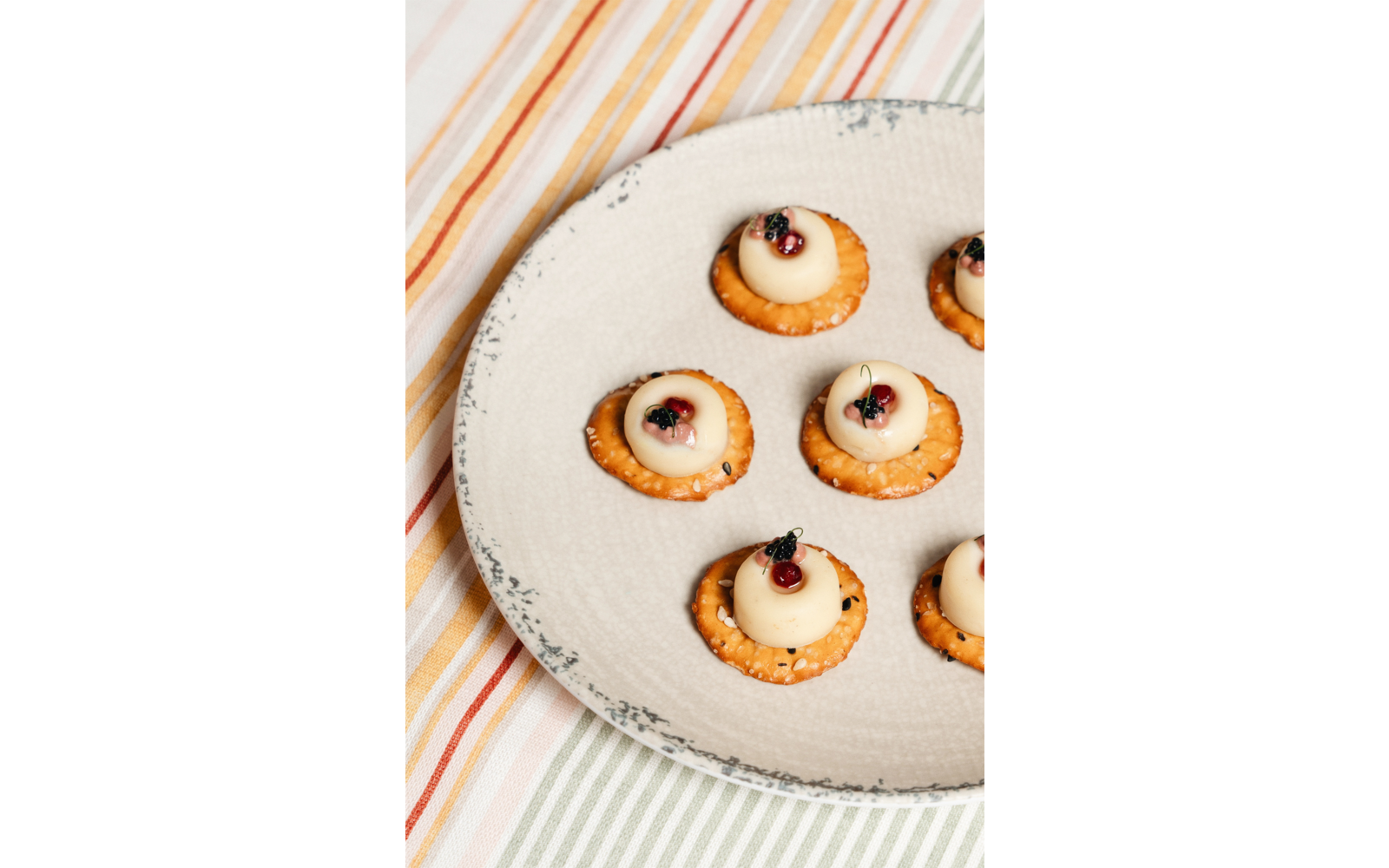 Sechs runde Cracker mit weißen Tupfen, darauf kleine rote Beeren und grüne Kräuterblättchen, auf einem beigen Teller auf einem gestreiften Tischtuch