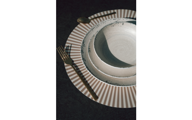 Drei ineinander gestapelte Teller auf dunklem Tuch: ein großer Teller mit braun-weißen Strahlen am Rand, zwei kleinere helle Teller mit dunklem, leicht unregelmäßigem Rand; goldfarbene Gabel links, goldfarbener Löffel oben