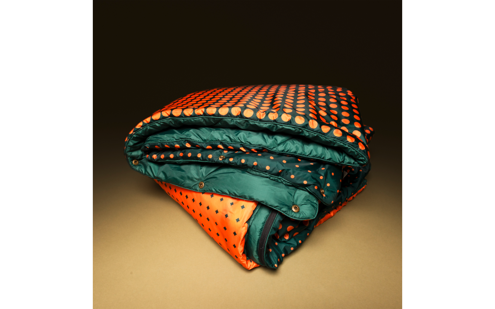 Folded quilt with orange polka-dot outer fabric and dark green inner lining with orange dots, visible snap buttons, on a brown background