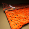 Two overlapping textiles on a beige surface; top fabric dark with orange dots, beneath a bright orange fabric with small black squares; visible folds and a partial white print