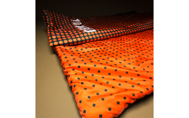 Two overlapping textiles on a beige surface; top fabric dark with orange dots, beneath a bright orange fabric with small black squares; visible folds and a partial white print