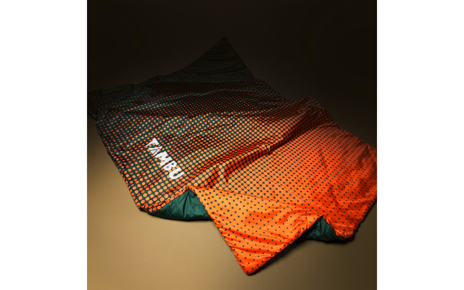 Spread-out orange sleeping bag with black dots, green inner lining and white brand text "TAMBU" on a beige background