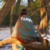Person sitting with back to the camera on a fallen tree branch, wrapped in a dark green blanket with orange polka dots and the white word 'TAMBO'; sandy ground and trees in the background