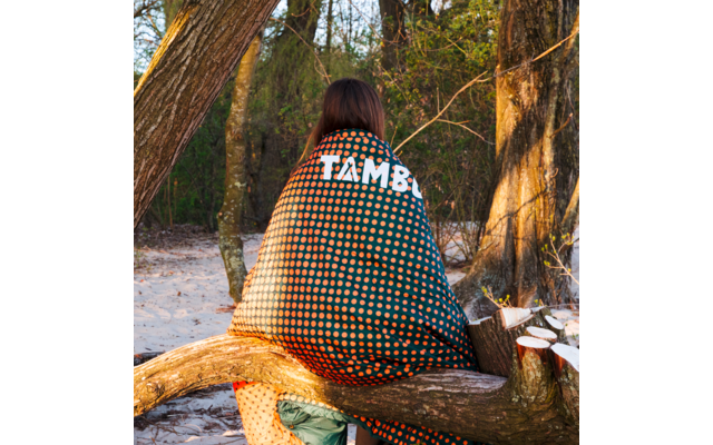 Person sitting with back to the camera on a fallen tree branch, wrapped in a dark green blanket with orange polka dots and the white word 'TAMBO'; sandy ground and trees in the background