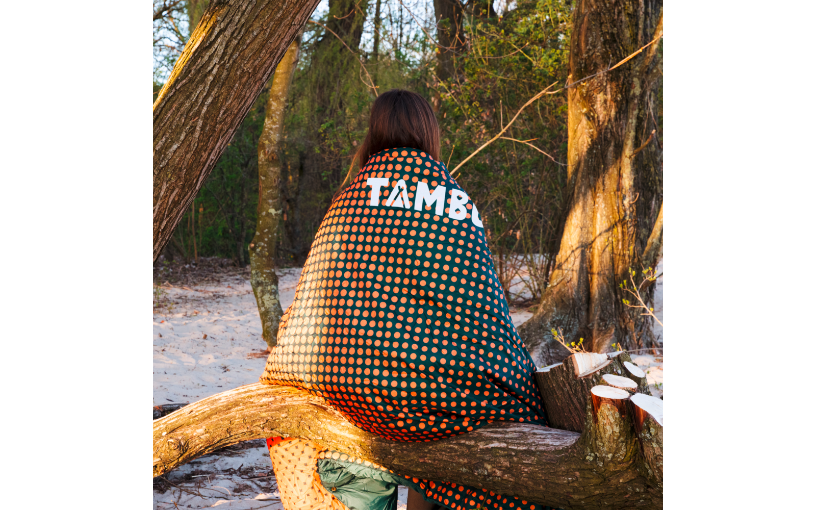 Person sitting with back to the camera on a fallen tree branch, wrapped in a dark green blanket with orange polka dots and the white word 'TAMBO'; sandy ground and trees in the background
