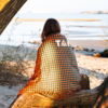 Person with shoulder-length brown hair sits on a tree trunk on a sandy beach, wrapped in an orange-and-black polka-dot blanket with white letters; sea and dunes in the background
