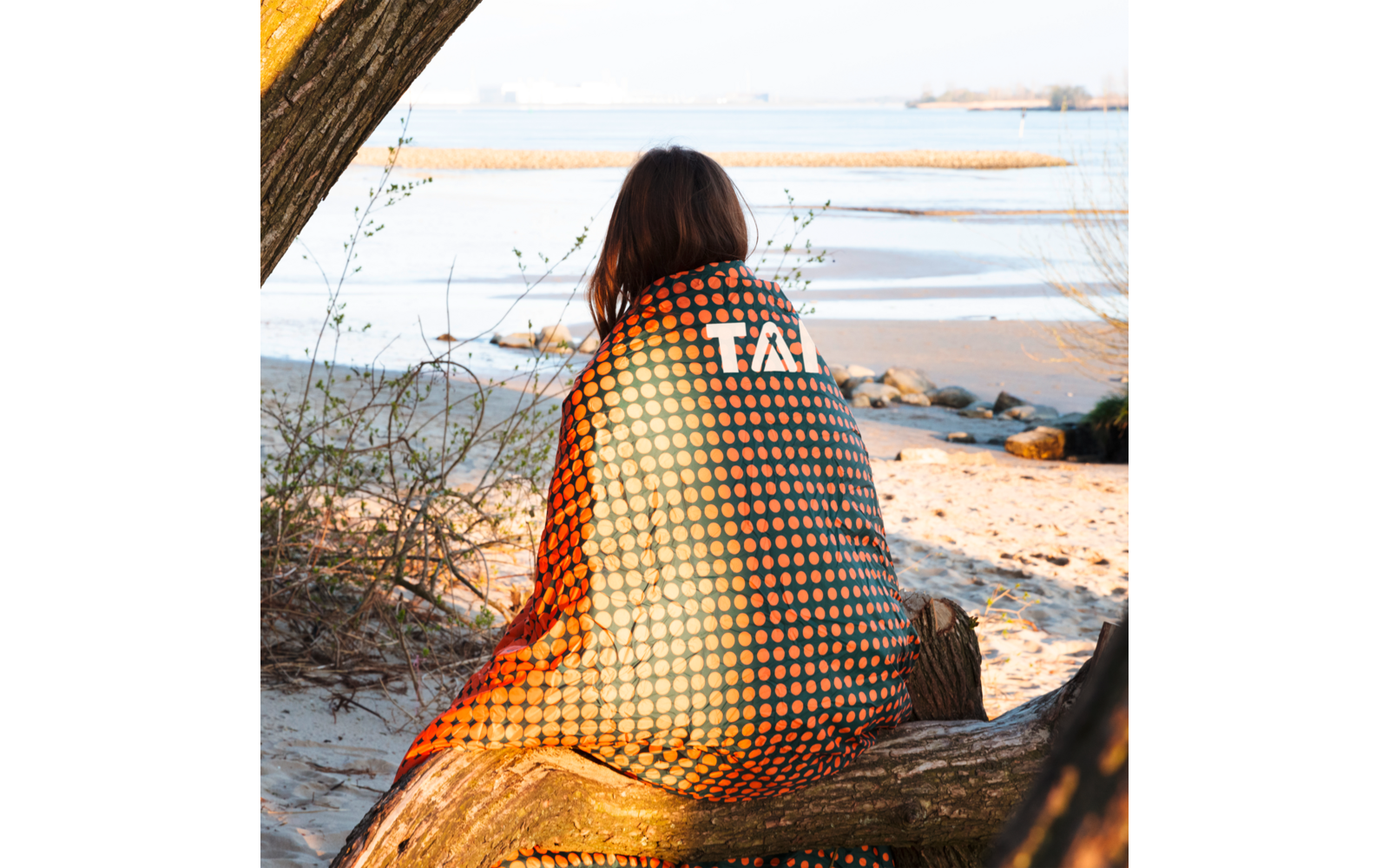 Person with shoulder-length brown hair sits on a tree trunk on a sandy beach, wrapped in an orange-and-black polka-dot blanket with white letters; sea and dunes in the background