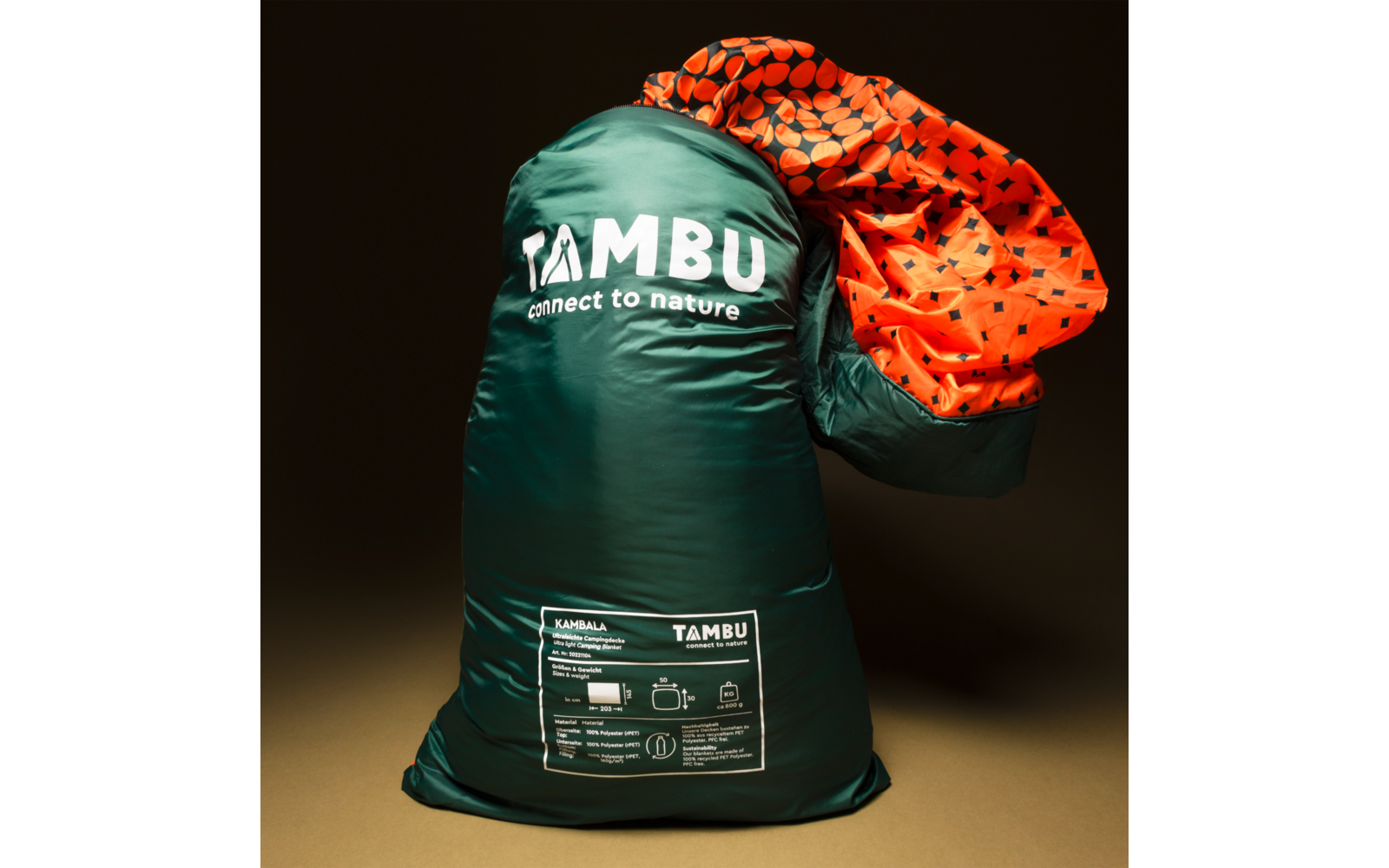 Dark green stuff sack with white "TAMBU connect to nature" logo and white product label at the bottom; orange patterned fabric protrudes from the opening; dark background