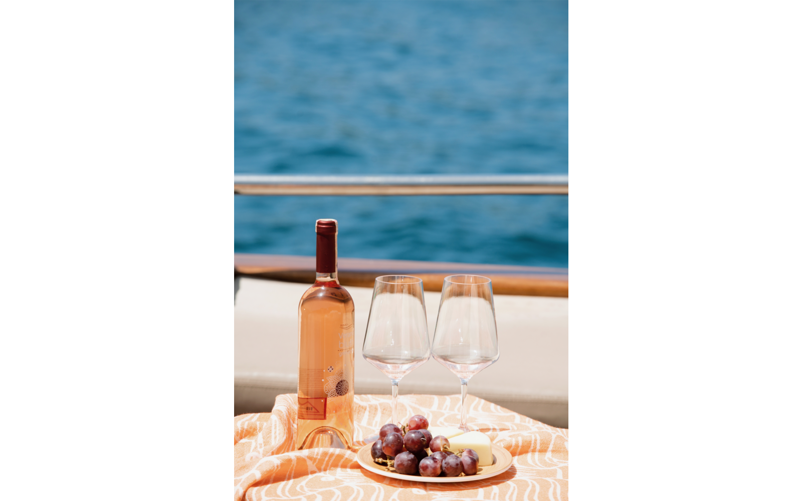 Flasche Roséwein, zwei leere Weingläser und ein Teller mit Trauben und Käse auf einem gemusterten Tuch, Reling und Meer im Hintergrund