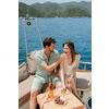 Zwei Personen sitzen auf dem Deck eines Boots; eine füttert die andere. Auf dem Tisch eine Roséflasche, Trauben und ein Teller; Meer und Berge im Hintergrund