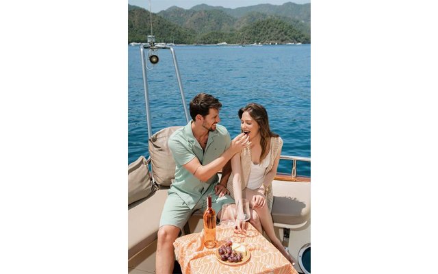 Zwei Personen sitzen auf dem Deck eines Boots; eine füttert die andere. Auf dem Tisch eine Roséflasche, Trauben und ein Teller; Meer und Berge im Hintergrund