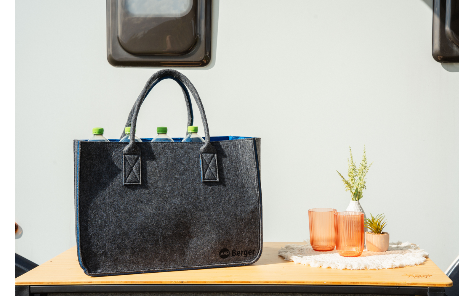 Graue Filztasche mit zwei Henkeln auf einer Holzfläche, aus der grüne Flaschenverschlüsse ragen; daneben zwei orangefarbene Gläser und zwei kleine Topfpflanzen auf einem weißen Tuch; heller Hintergrund mit zwei kleinen Fenstern