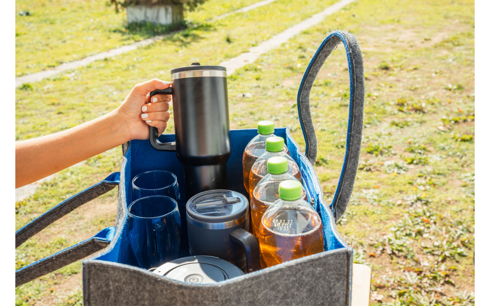 Hand hält Thermoskanne über einer grauen Stofftasche mit blauer Innenfläche; in der Tasche stehen mehrere Plastikflaschen, Becher und Behälter; Gras im Hintergrund