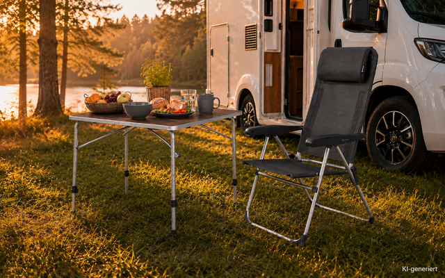 Weißer Camper mit geöffneter Tür auf einer Wiese am See; Klapptisch mit Schalen, Karaffe und Tassen; schwarzer Campingstuhl; Bäume und Wasser im Hintergrund