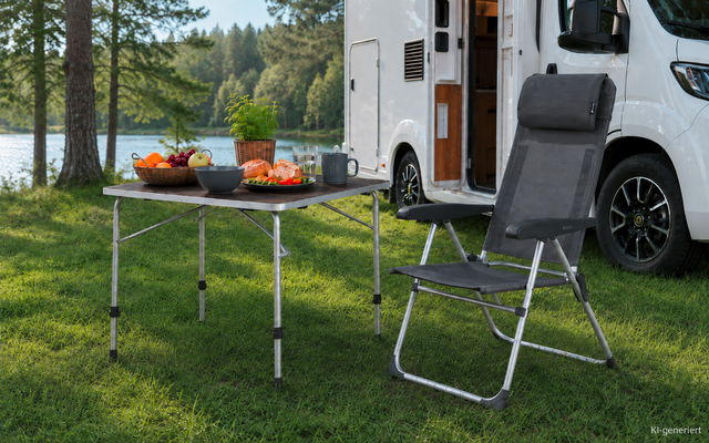 Camper geparkeerd bij een meer, daarvoor een inklapbare tafel met kommen met fruit en brood, naast de tafel een opvouwbare campingstoel, gras en bomen op de achtergrond
