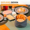 Table avec petit-déjeuner : croissants dans un panier en tissu, un petit pain sur une assiette, tranches de fromage et de charcuterie, deux œufs dans des coquetiers, un morceau de fromage et deux bouteilles ; en haut le texte « ZEITLOSES DESIGN » et « PERFEKT FÜR FRÜHSTÜCK & ABENDBROT »