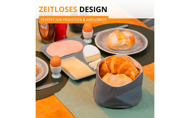 Table avec petit-déjeuner : croissants dans un panier en tissu, un petit pain sur une assiette, tranches de fromage et de charcuterie, deux œufs dans des coquetiers, un morceau de fromage et deux bouteilles ; en haut le texte « ZEITLOSES DESIGN » et « PERFEKT FÜR FRÜHSTÜCK & ABENDBROT »