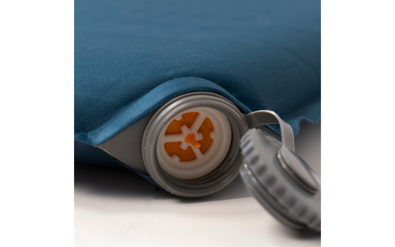 Nahaufnahme eines blauen Stoffteils mit grauem Kunststoffventil, orangefarbene Innenkomponenten, abgelegter Ventildeckel daneben