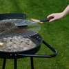 Main tenant une brosse à gril au-dessus d'un barbecue rond noir au charbon avec une grille métallique; cendres et charbon à l'intérieur, gazon en arrière-plan