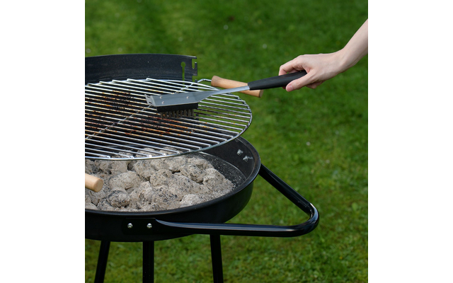 Main tenant une brosse à gril au-dessus d'un barbecue rond noir au charbon avec une grille métallique; cendres et charbon à l'intérieur, gazon en arrière-plan