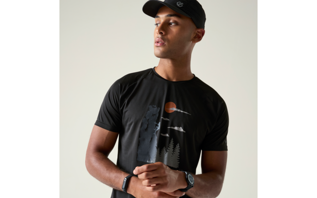 Person mit schwarzer Kappe und schwarzem T‑Shirt mit Wald- und Sonnenprint, schaut nach rechts, Hände vor dem Bauch gefaltet, Smartwatch am Handgelenk