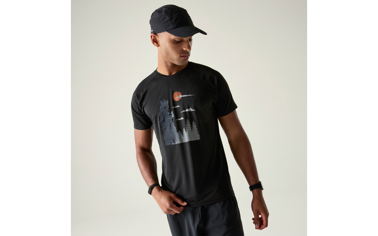 Mann mit schwarzer Kappe und schwarzem T‑Shirt mit Motiv (stehender Bär, Tannen, Vögel, orangene Sonne); schaut nach unten, Hände an der Hüfte, heller Hintergrund