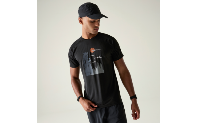 Mann mit schwarzer Kappe und schwarzem T‑Shirt mit Motiv (stehender Bär, Tannen, Vögel, orangene Sonne); schaut nach unten, Hände an der Hüfte, heller Hintergrund