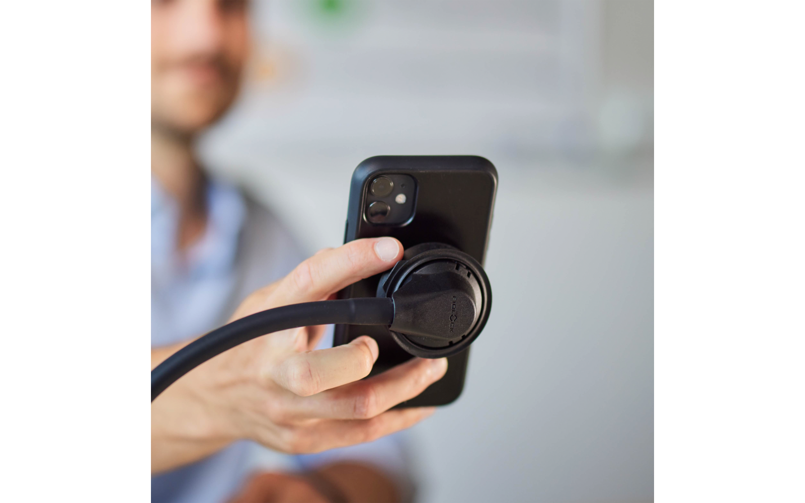 Hand hält ein Smartphone; Stethoskop-Bruststück liegt auf der Rückseite des Telefons; unscharfe Person im Hintergrund
