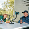Zwei Personen sitzen an einem Campingtisch neben einem Wohnmobil; an der Außensteckdose hängt ein Stromkabel. Auf dem Tisch Thermosflasche und Teller mit Gebäck, im Hintergrund Fahrräder und Bäume