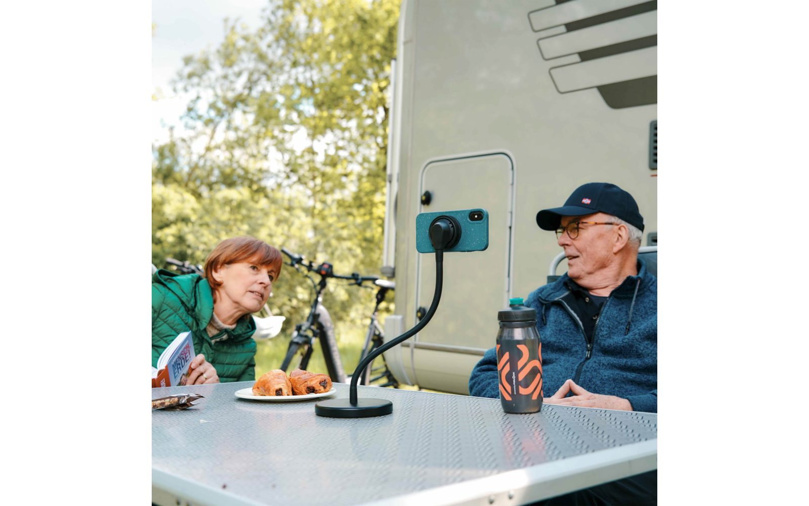 Zwei Personen sitzen an einem Campingtisch neben einem Wohnmobil; an der Außensteckdose hängt ein Stromkabel. Auf dem Tisch Thermosflasche und Teller mit Gebäck, im Hintergrund Fahrräder und Bäume