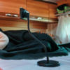 Flexibler Smartphone-Halter mit rundem Fuß auf einer Matratze; im Hintergrund schwarze Decke, gestreiftes Kissen, hölzerne Oberschränke und weißer Vorhang