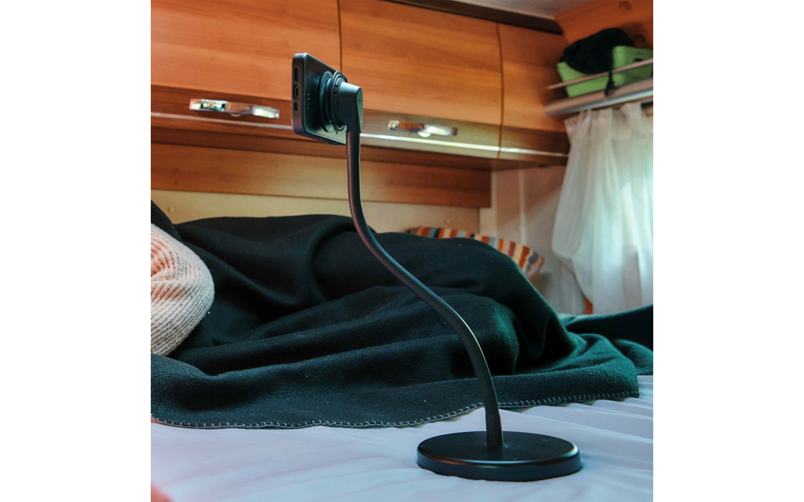 Flexibler Smartphone-Halter mit rundem Fuß auf einer Matratze; im Hintergrund schwarze Decke, gestreiftes Kissen, hölzerne Oberschränke und weißer Vorhang