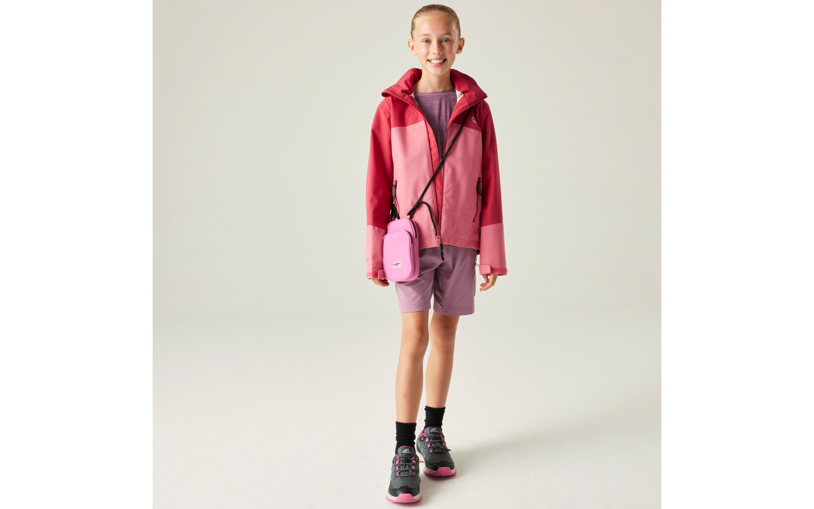 Person vor hellem Hintergrund, trägt rosa-rote Jacke, violette Shorts, schwarz-rosa Sneaker und kleine rosa Umhängetasche, lächelt