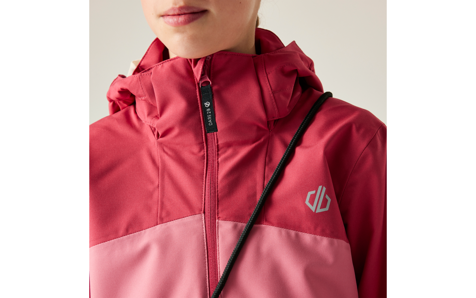 Unteres Gesicht (Mund und Kinn) einer Person, die eine zweifarbige rot-rosa Kapuzenjacke mit hochgezogenem Reißverschluss trägt; schwarzer Riemen diagonal über der Brust, kleines graues Logo auf der Jacke