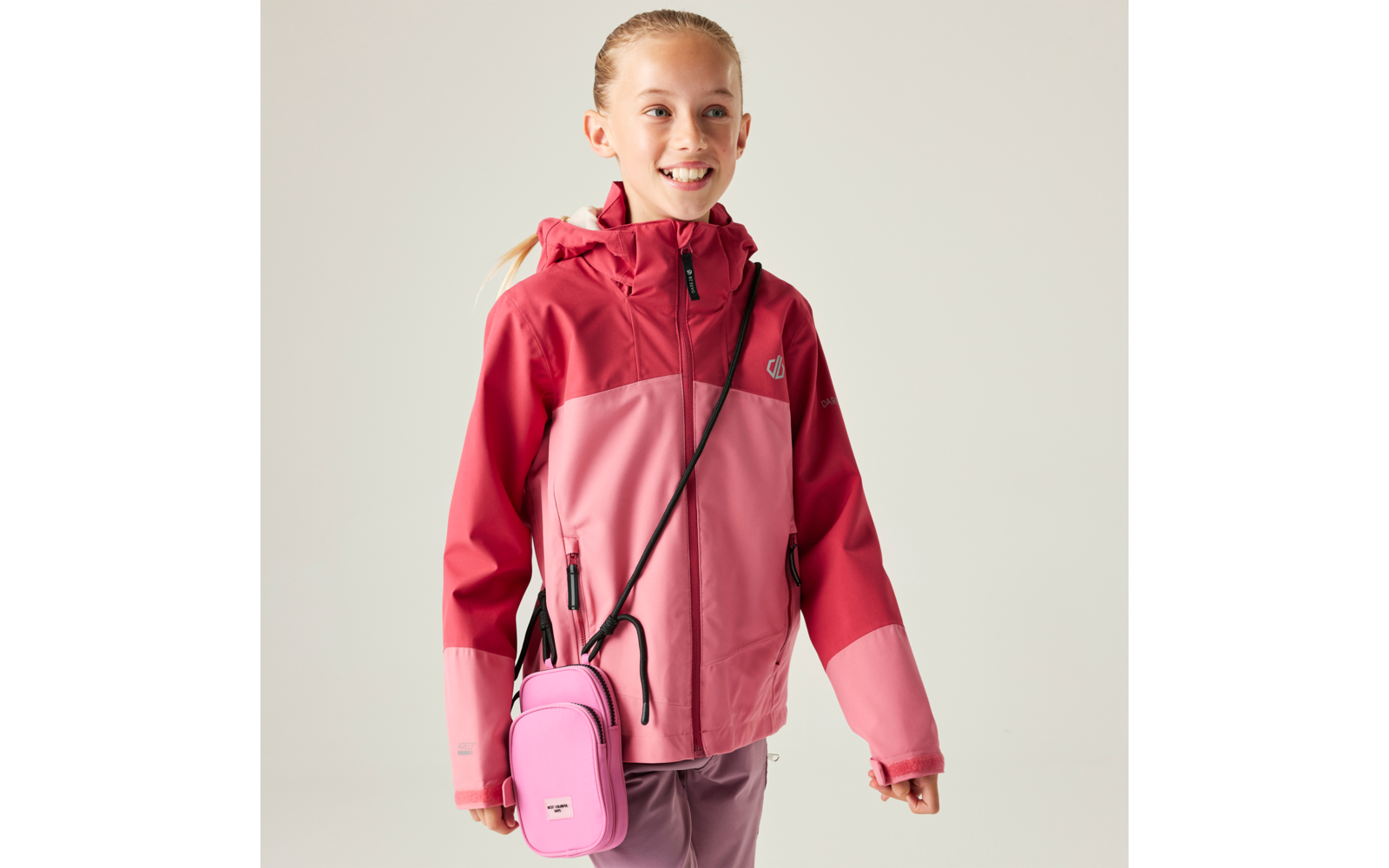 Mädchen mit Pferdeschwanz trägt zweifarbige rosa-rote Jacke mit Kapuze und kleine rosa Umhängetasche vor hellem Hintergrund