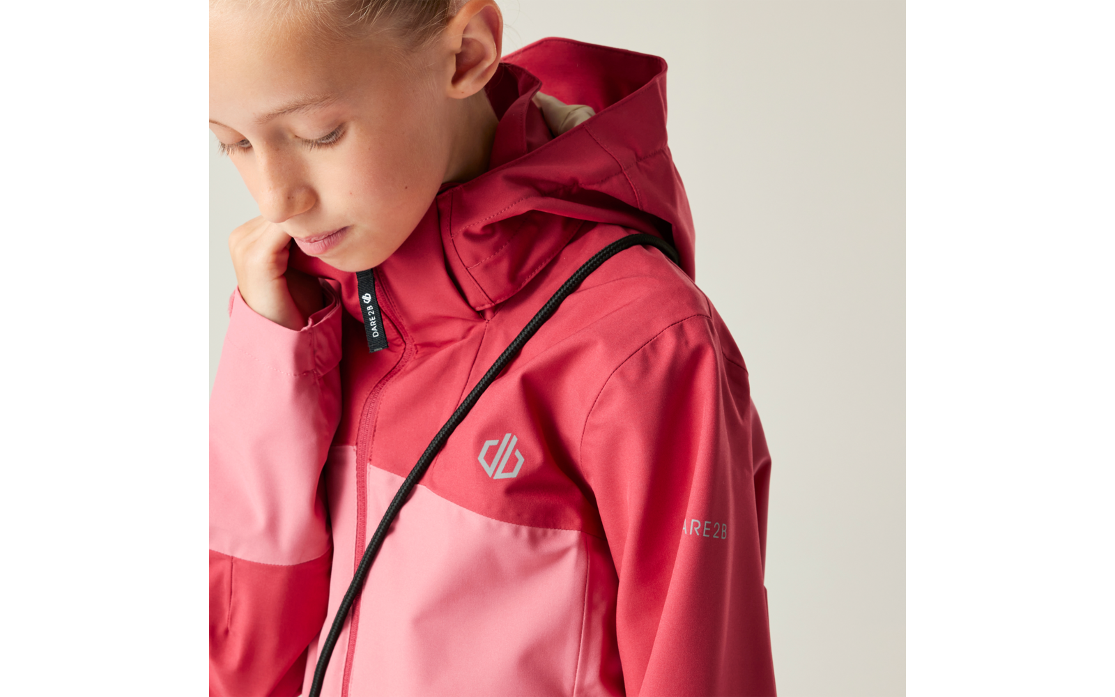 Kind mit zusammengebundenem blondem Haar, blickt nach unten, trägt rot-rosa Regenjacke mit Kapuze, schwarzem Kordelzug, Logo auf der Brust und Schriftzug am Ärmel