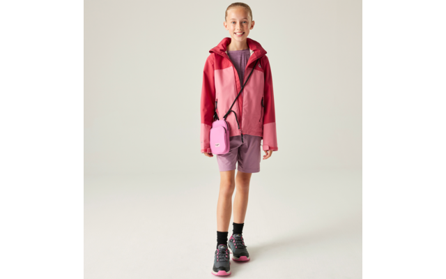 Person vor hellem Hintergrund, trägt rosa-rote Jacke, violette Shorts, schwarz-rosa Sneaker und kleine rosa Umhängetasche, lächelt