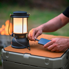 Lanterna da campeggio nera su una cassa di plastica; due mani tengono uno smartphone e avvicinano un cavo alla lanterna; falò sfocato e erba sullo sfondo